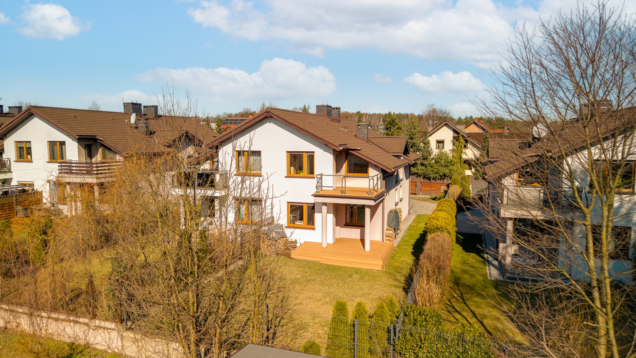 Dwupiętrowy dom z balkonem i tarasem z drewnianą podłogą, otoczony zielenią i innymi budynkami. Ujęcie z góry, widoczne detale architektury i otoczenia.