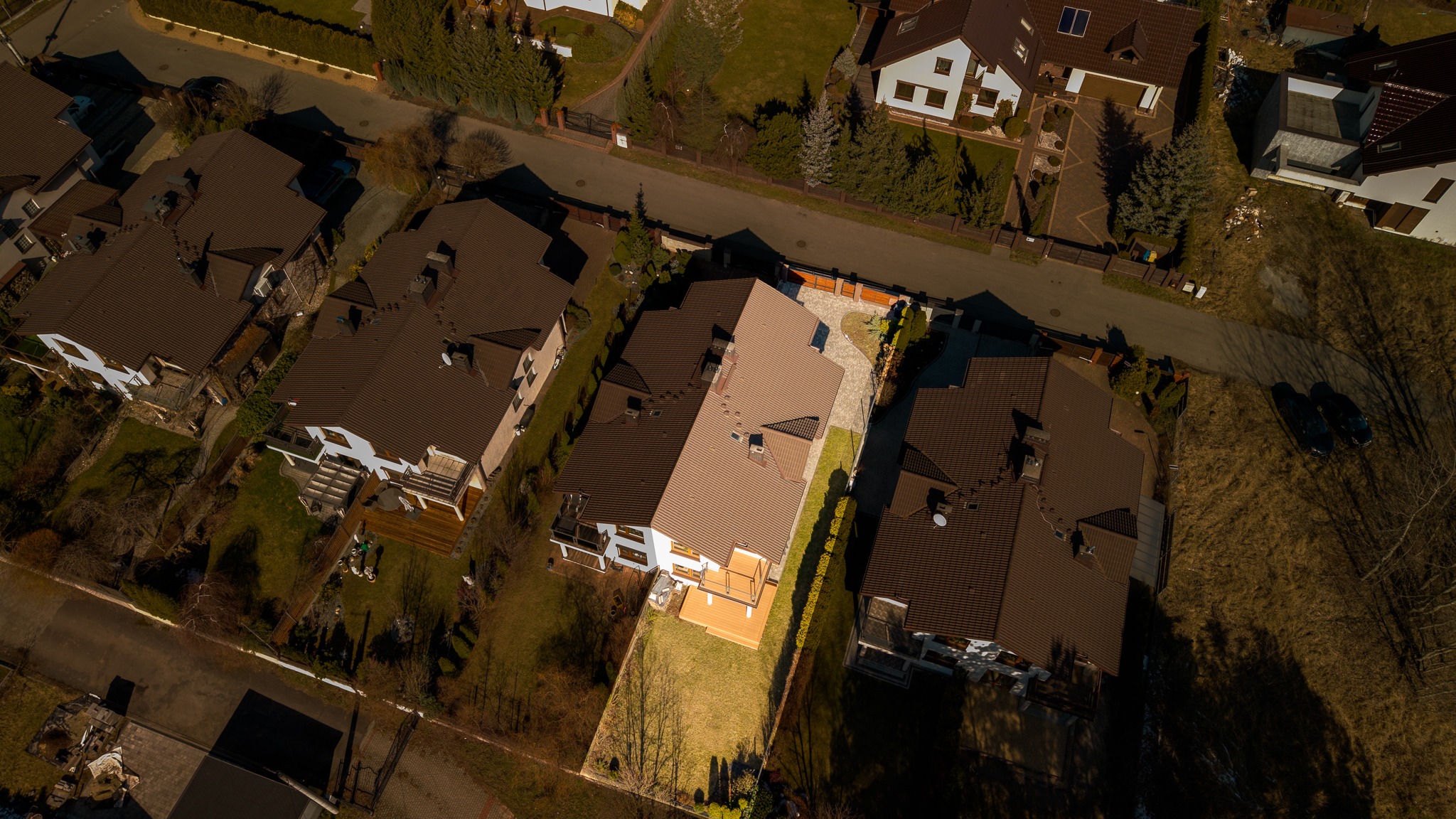 Zdjęcie z drona przedstawia osiedle domów jednorodzinnych z brązowymi dachami, otoczonych zielenią. Widoczna ulica i zadbane ogrody.