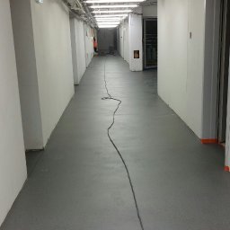 BW Floor Ewa Wołowicz - Długi korytarz z szarą, gładką posadzką. Na suficie widoczne rury i oświetlenie. Kabel elektryczny biegnie przez środek podłogi. Widok w perspektywie.