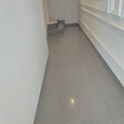 Jasnoszara, gładka posadzka z delikatnym, białym wzorem, widoczne schody w tle i dekoracyjne listwy przypodłogowe.