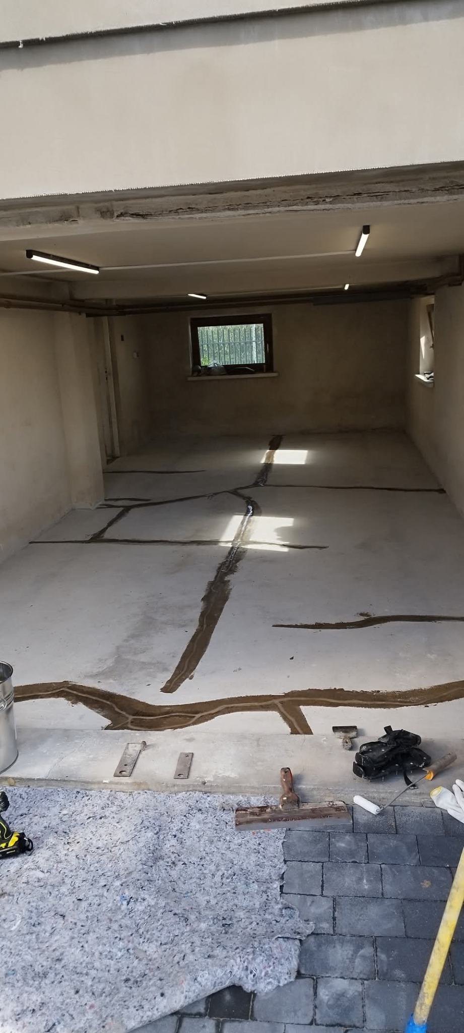 Przygotowanie podłoża pod posadzkę: wypełnione szczeliny, narzędzia, mata izolacyjna i kostka brukowa w surowym wnętrzu z oknem i lampami.