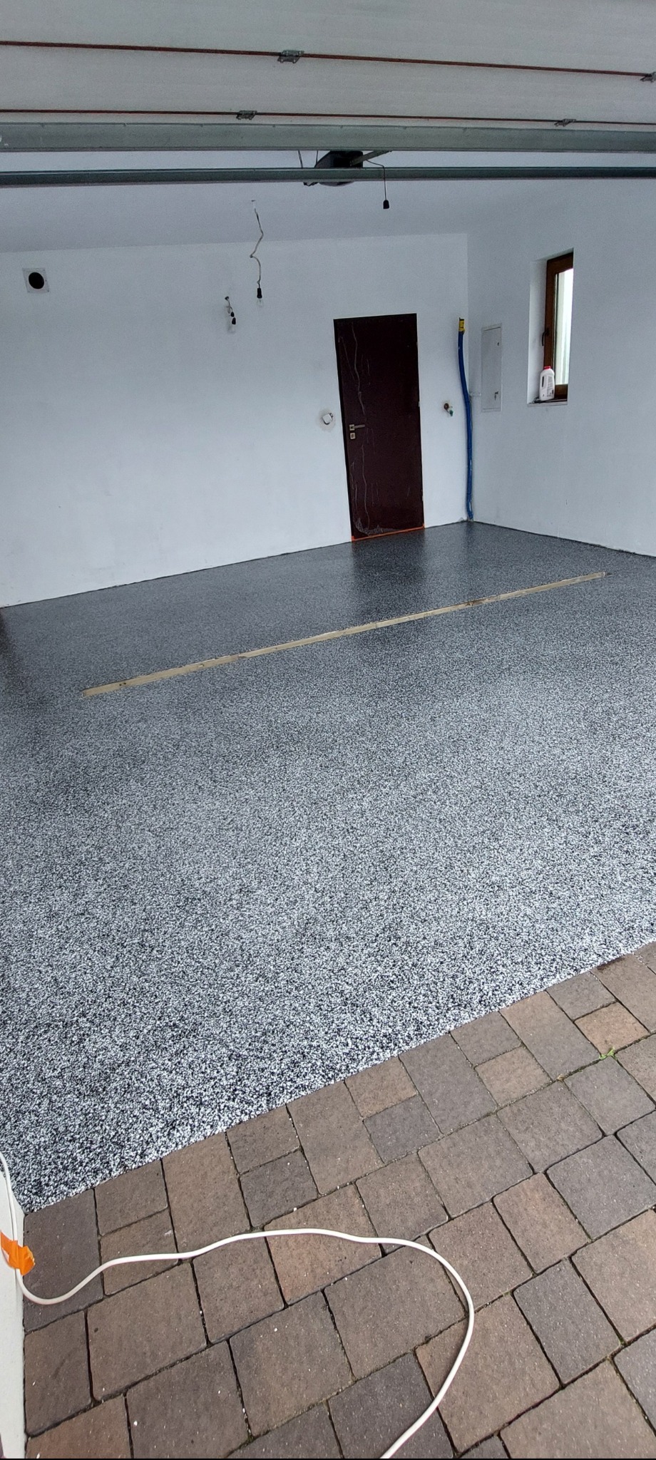 Szara posadzka żywiczna w garażu z widocznym łączeniem desek i przejściem na kostkę brukową. Na ścianie boczna brama i okno.