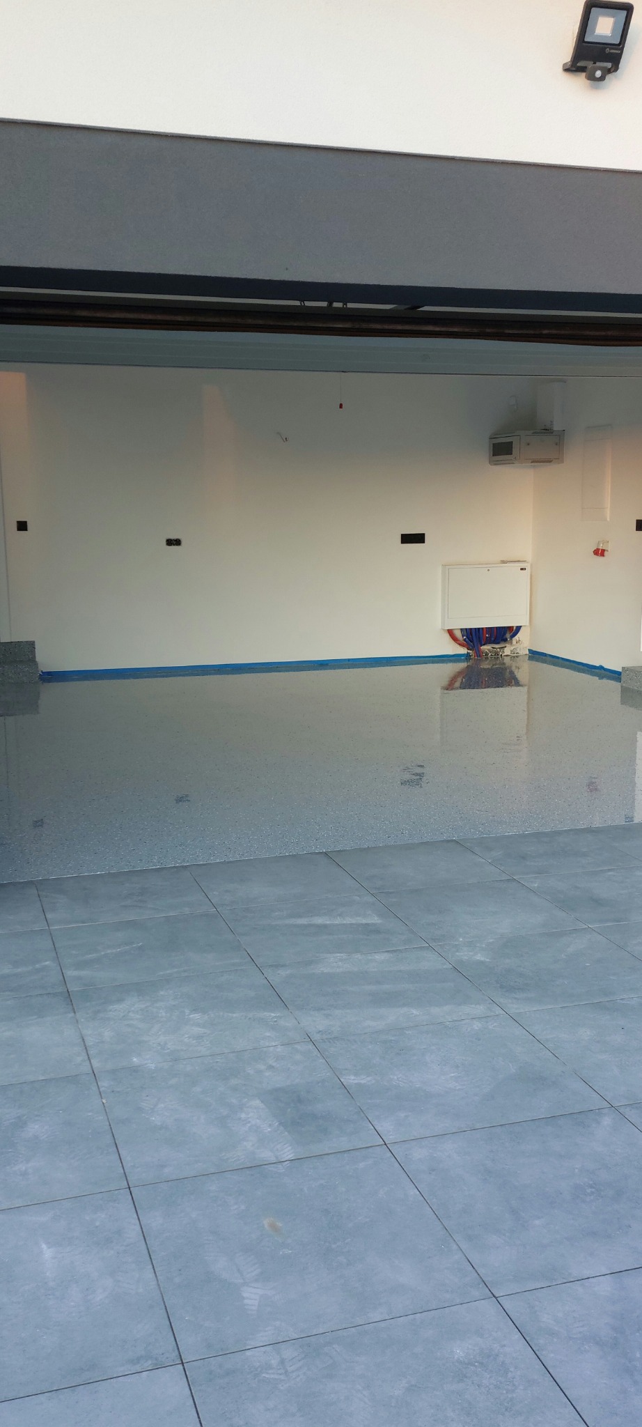 Wykończona, jasnoszara posadzka z delikatnym wzorem w garażu z białą ścianą i niebieską taśmą maskującą wzdłuż krawędzi. Widoczne płytki podłogowe w dolnej części kadru.