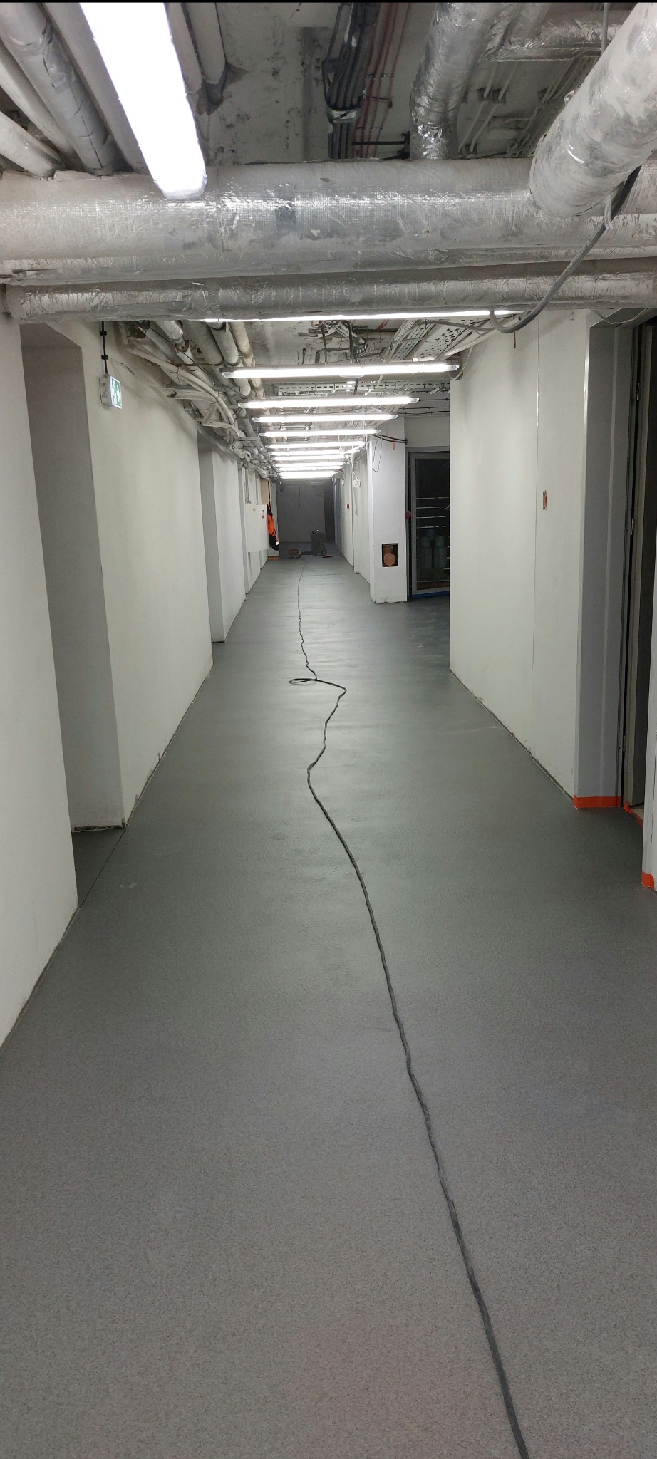Długi korytarz z szarą, gładką posadzką. Na suficie widoczne rury i oświetlenie. Kabel elektryczny biegnie przez środek podłogi. Widok w perspektywie.
