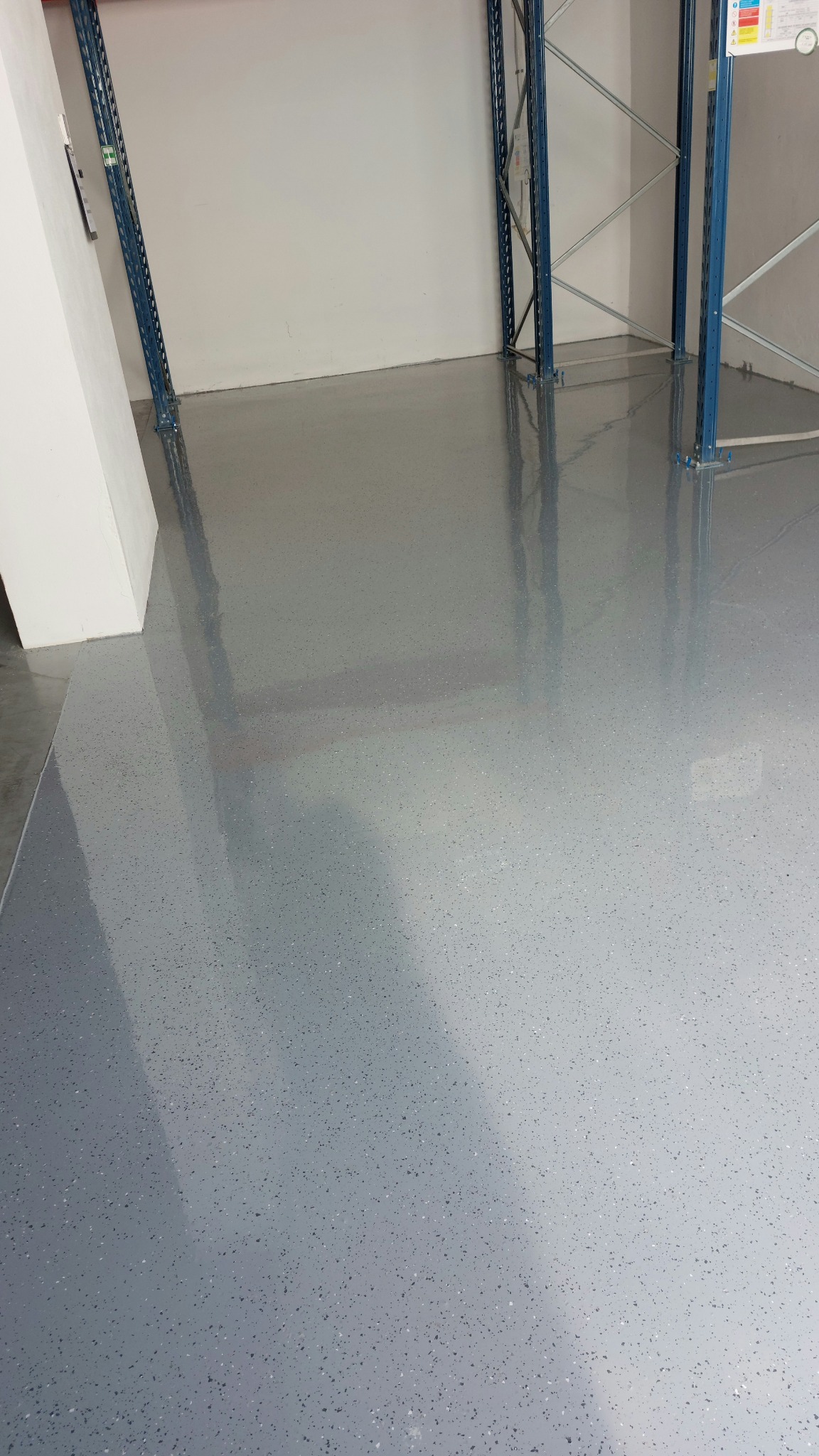 Gładka, szara posadzka z delikatnym wzorem w hali magazynowej, z widocznymi elementami regałów. Powierzchnia odbija światło, podkreślając równomierne wykończenie.