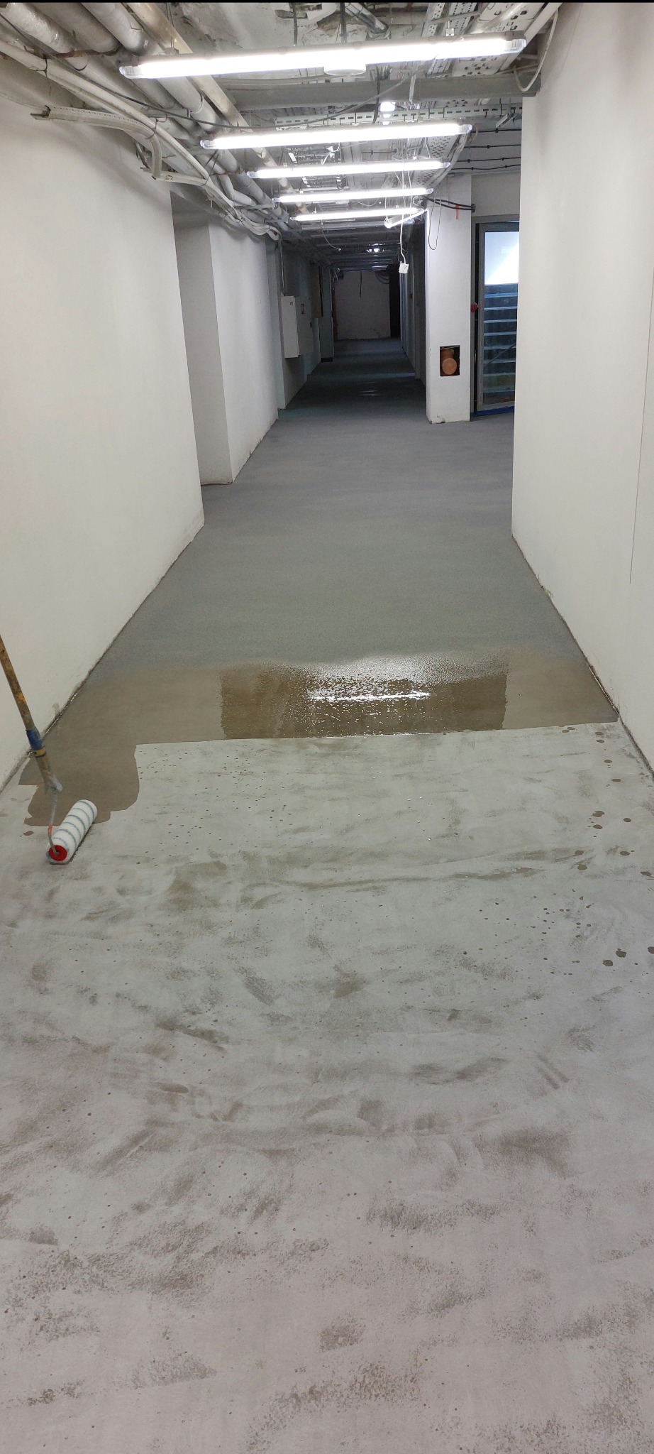 Aplikacja posadzki w korytarzu. Częściowo mokra powierzchnia, wałek malarski przy ścianie. Widoczne rury pod sufitem. Oświetlenie fluorescencyjne.