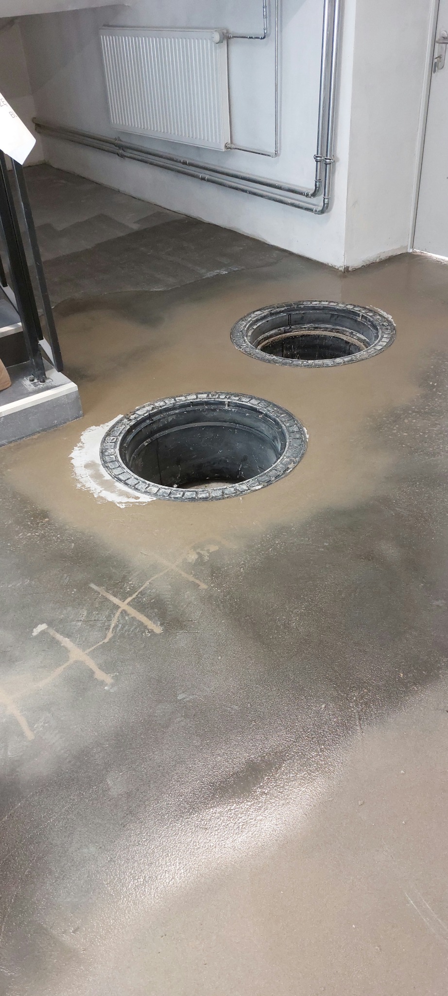 Szara, błyszcząca posadzka z dwoma okrągłymi otworami rewizyjnymi. Widoczne ślady po wyznaczeniu geometrii podłoża. W tle ściana z rurami i grzejnikiem.