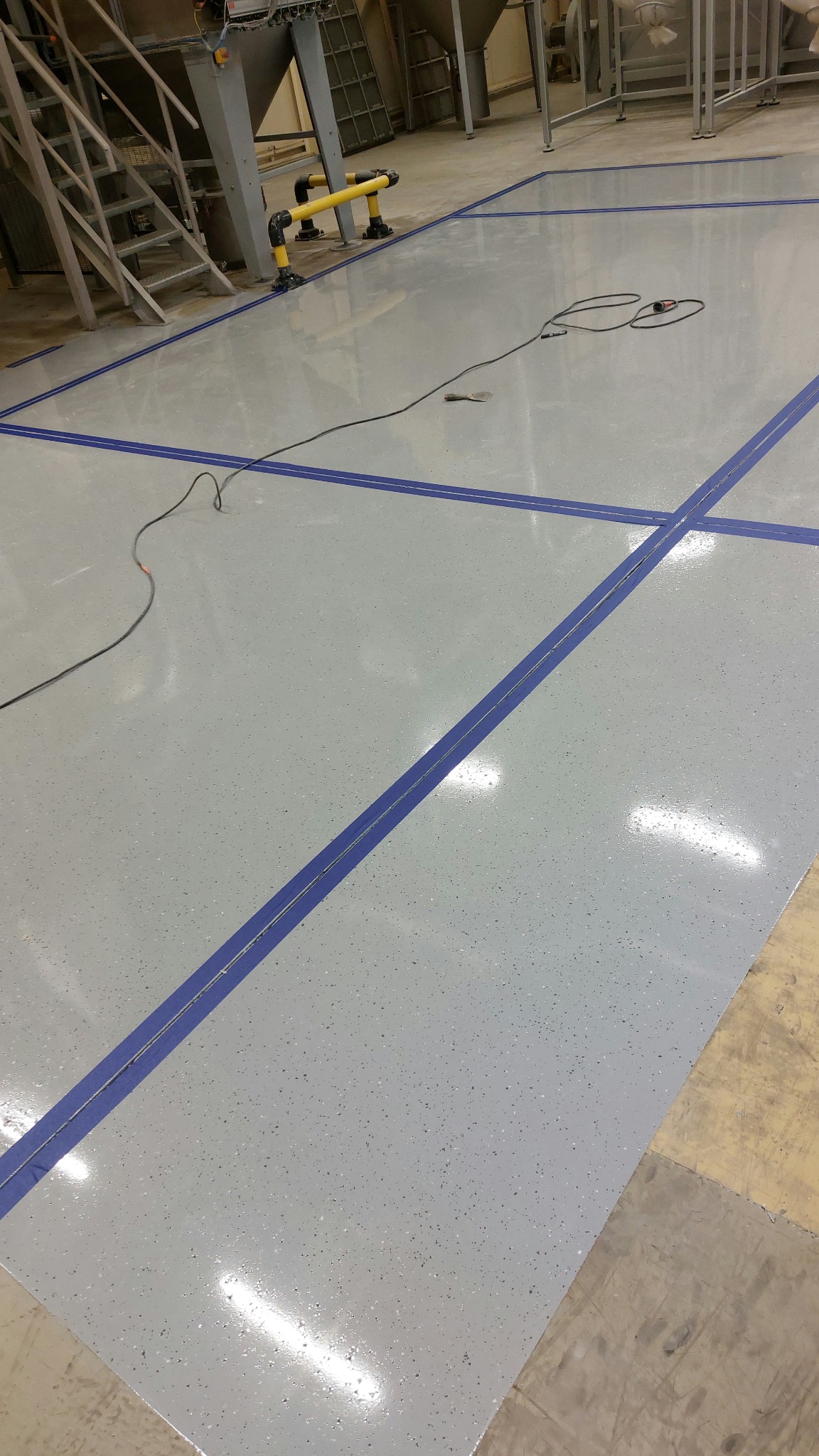 Jasnoszara posadzka przemysłowa z niebieskimi liniami podziału, narzędzia i schody w tle, widok z góry, oświetlenie górne.