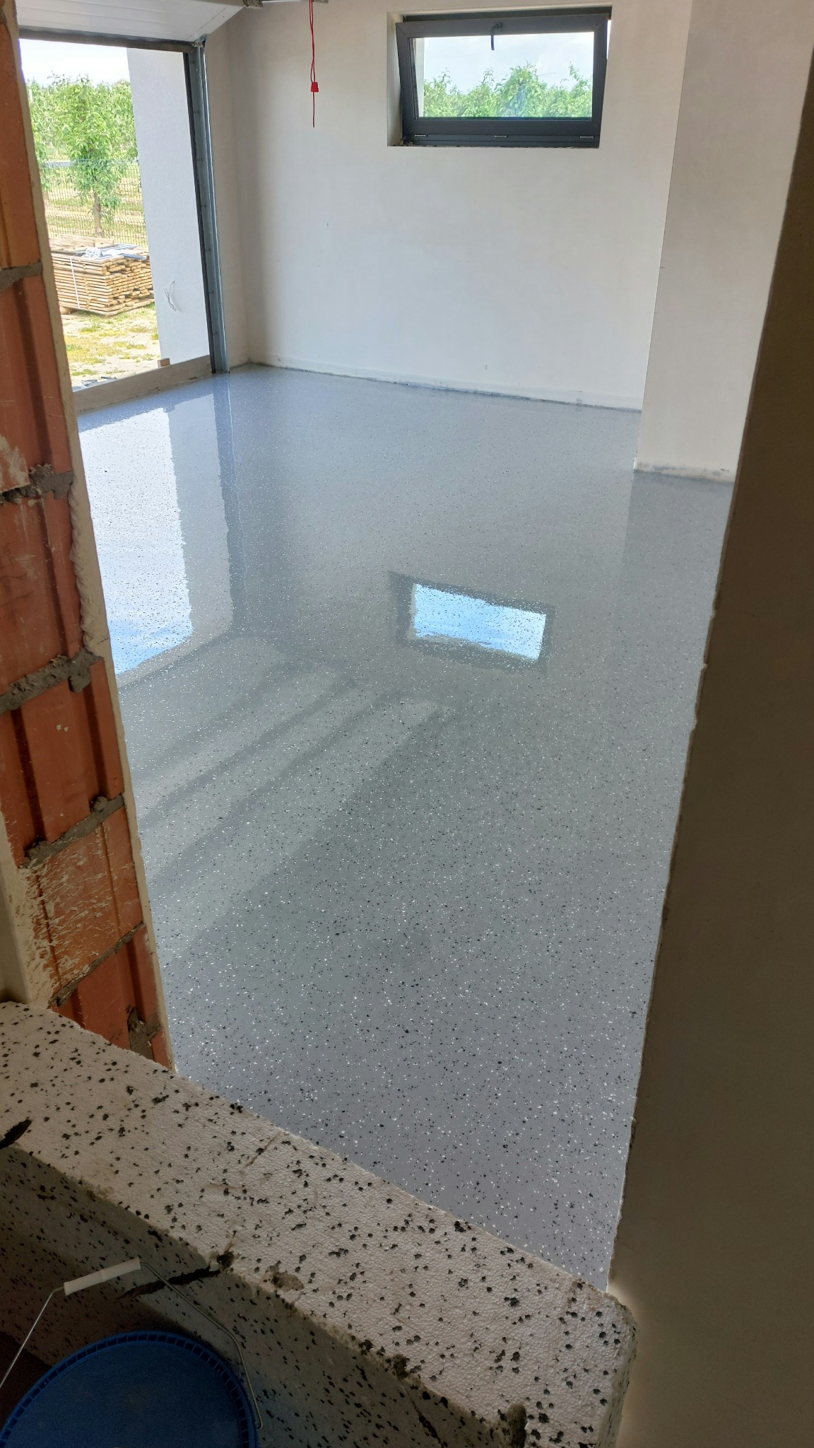 Świeżo wylana, błyszcząca, szara posadzka żywiczna z widocznymi drobinkami, odbijająca okno. Surowy stan budynku. Kadr z perspektywy wejścia.