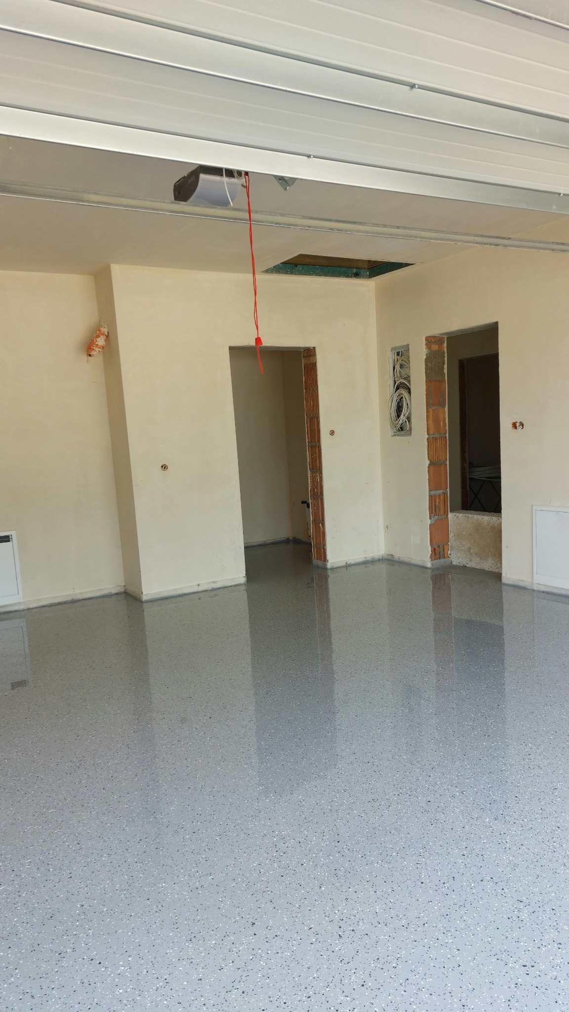 Błyszcząca, szara posadzka z drobnymi plamkami w surowym wnętrzu z widocznymi instalacjami i otworami w ścianach. Nowoczesna realizacja w trakcie wykończenia.