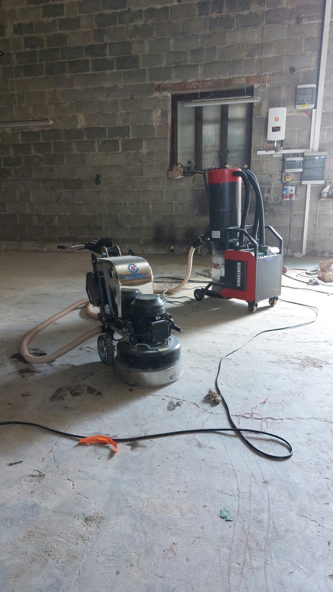 Maszyny do szlifowania posadzek na betonowej podłodze w surowym wnętrzu z ceglaną ścianą i oknem. Widoczne kable i węże.