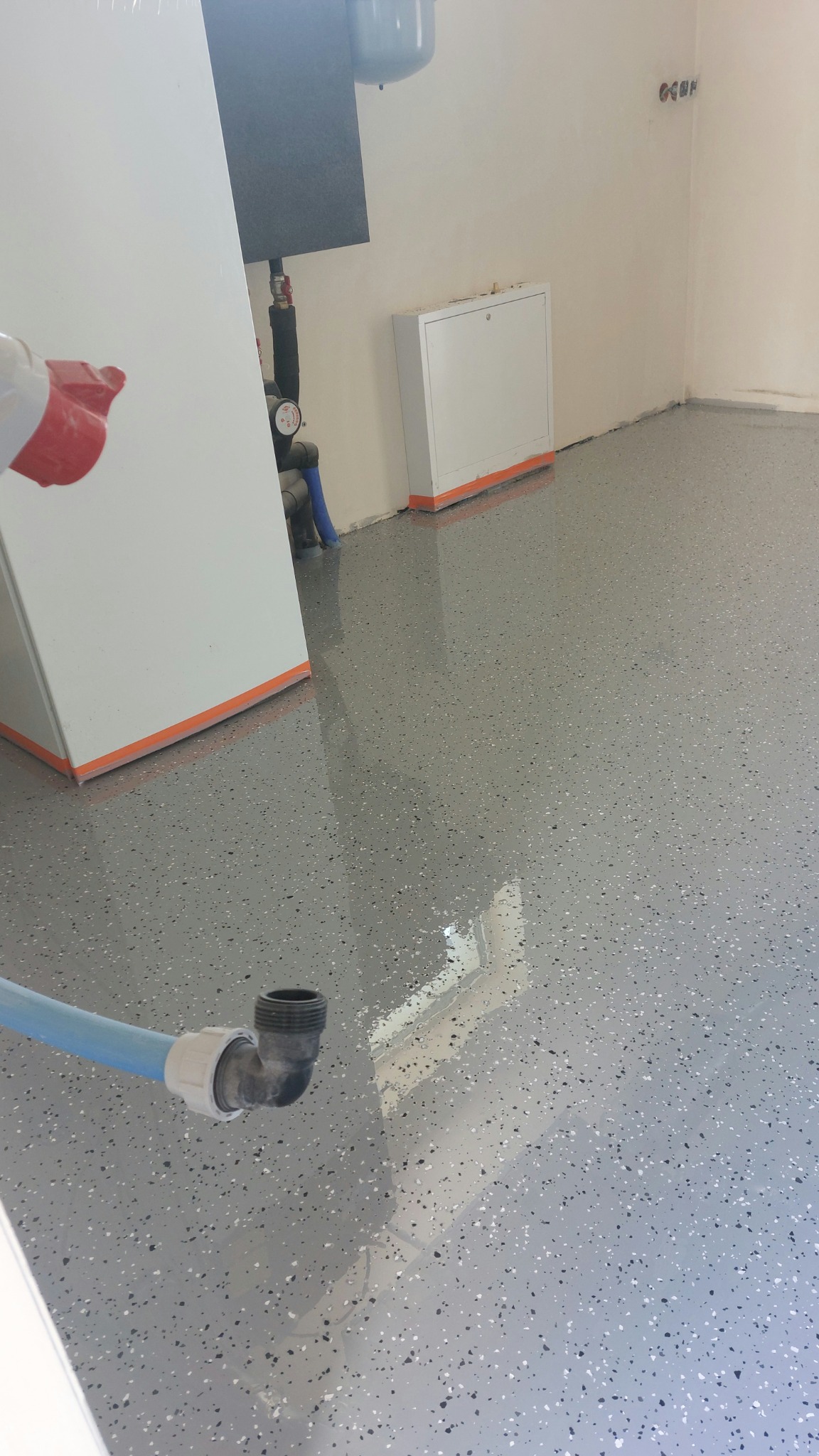 Błyszcząca, szara posadzka żywiczna z białymi i czarnymi drobinkami w pomieszczeniu z białą szafką i instalacją grzewczą. Widoczny fragment rury.