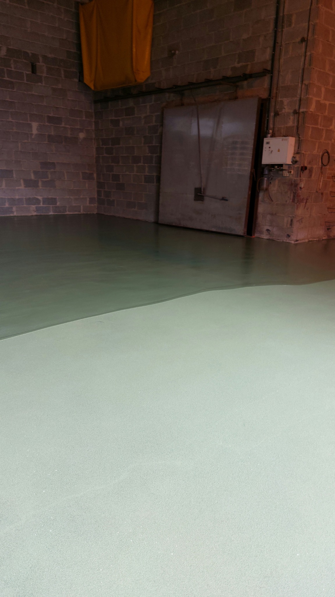 Dwuwarstwowa, gładka posadzka w hali produkcyjnej: dolna warstwa jasna, górna oliwkowa, na tle ściany z szarego pustaka i metalowych drzwi.