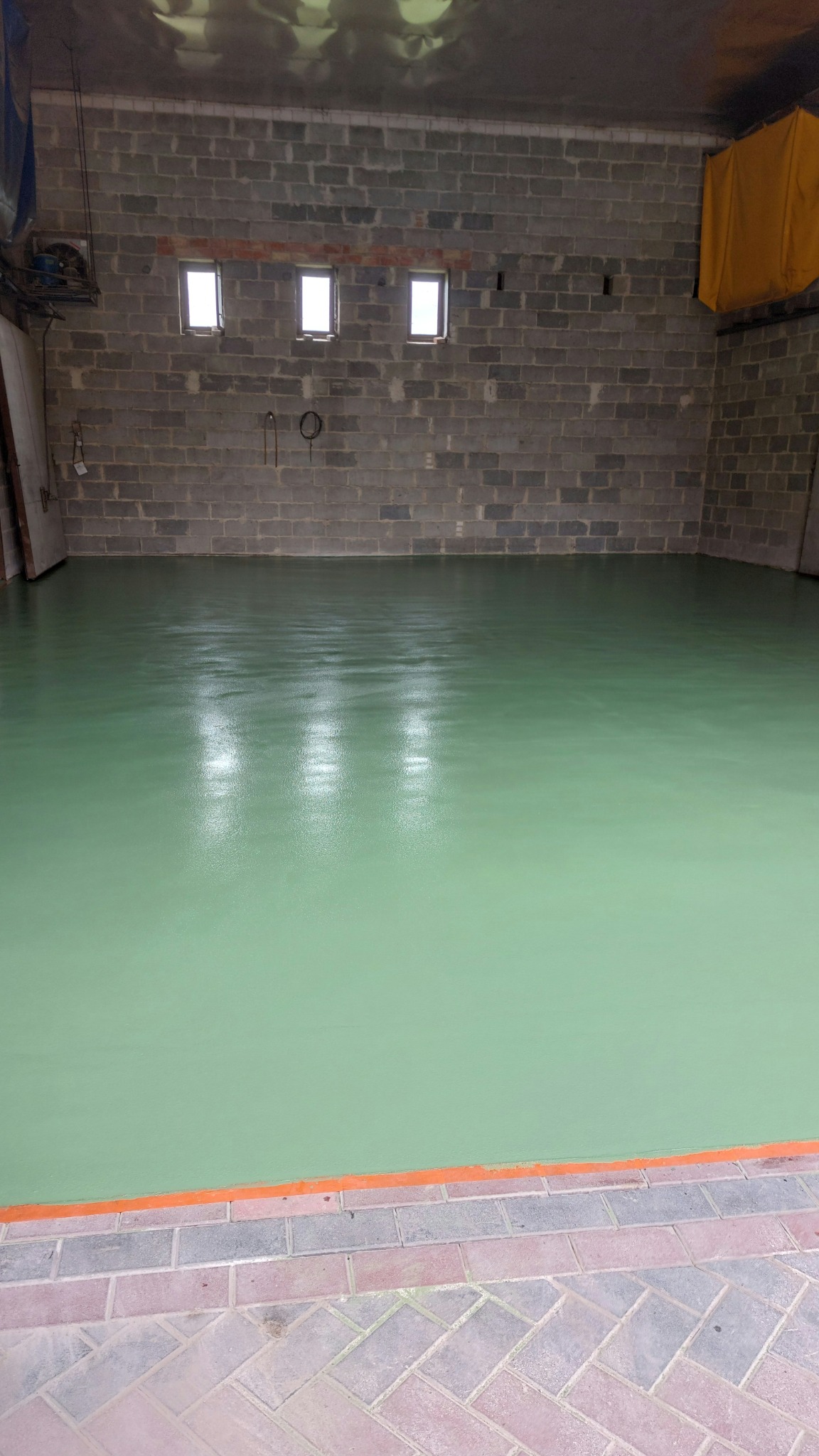 Jasnozielona, błyszcząca posadzka w garażu z szarego betonu komórkowego. Trzy małe okna w ścianie. Pomarańczowa listwa przy podłodze.