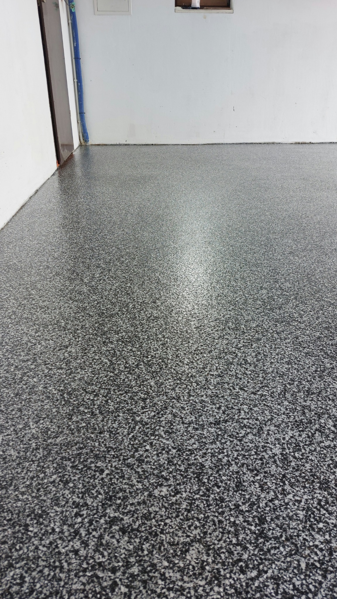 Szara, błyszcząca posadzka z drobnymi, białymi i czarnymi elementami, wykonana w pomieszczeniu z białymi ścianami. Widoczny fragment ściany i rury.