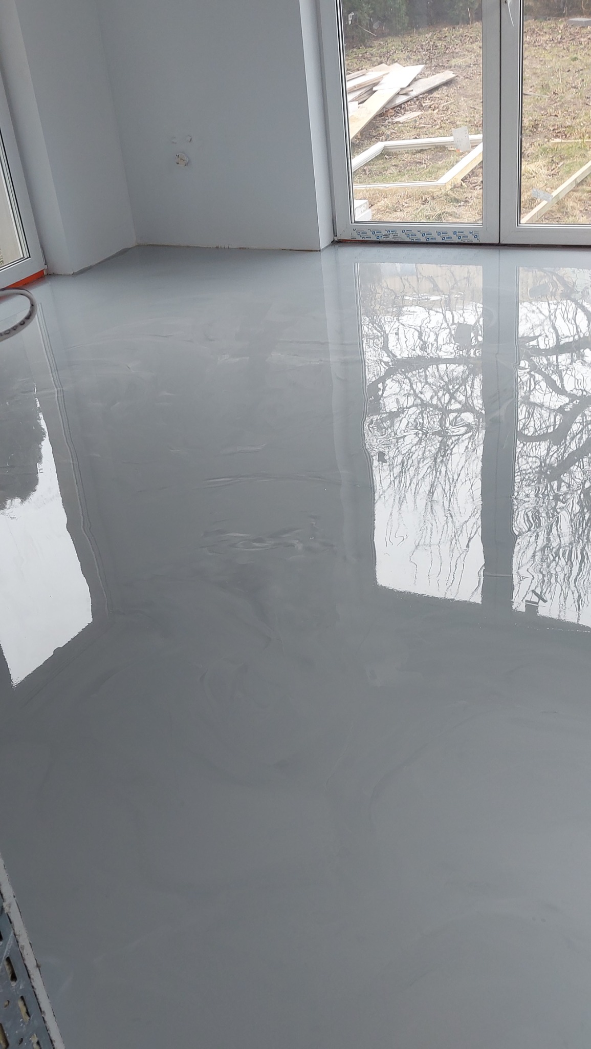 Świeżo wylana, gładka, szara posadzka w jasnym pomieszczeniu z dużymi oknami, w których odbija się drzewo. Widoczne detale wykończenia.