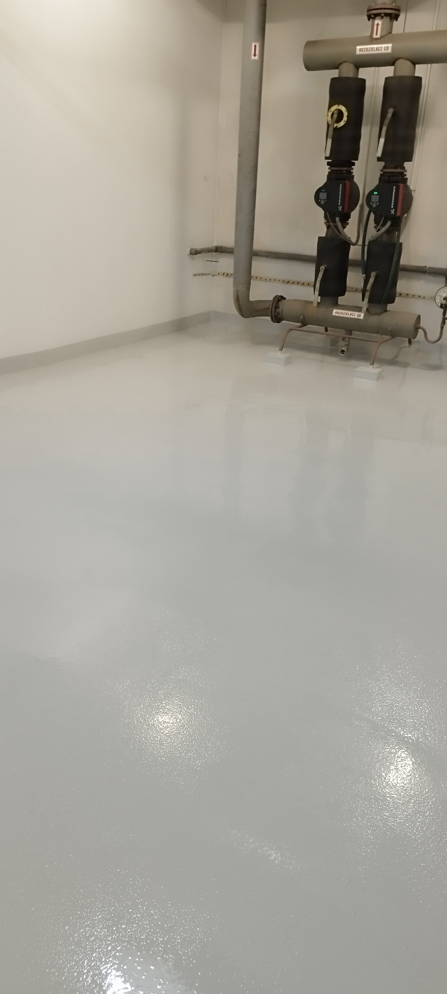 Gładka, jasnoszara posadzka w pomieszczeniu technicznym z rurami i elementami instalacji grzewczej. Błyszcząca powierzchnia, widoczne refleksy świetlne.