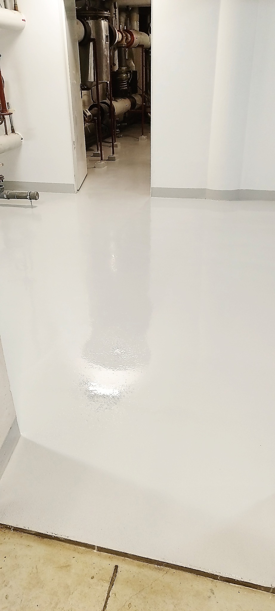 Biała, lśniąca posadzka żywiczna w pomieszczeniu technicznym z widocznymi rurami i instalacjami. Gładka powierzchnia, jasne oświetlenie, widoczny fragment surowej podłogi.