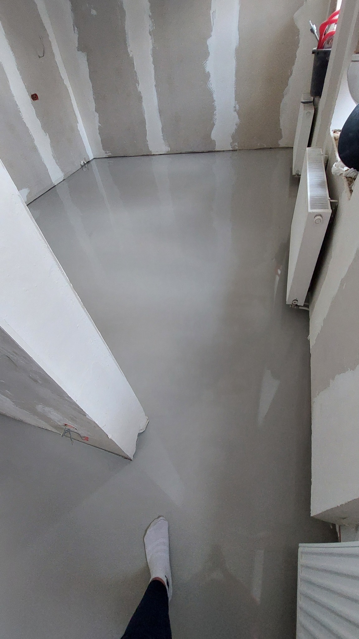 Świeżo wylana, gładka posadzka w szarym odcieniu w pomieszczeniu w trakcie remontu. Widoczne ślady po taśmach na ścianach i fragment grzejnika.