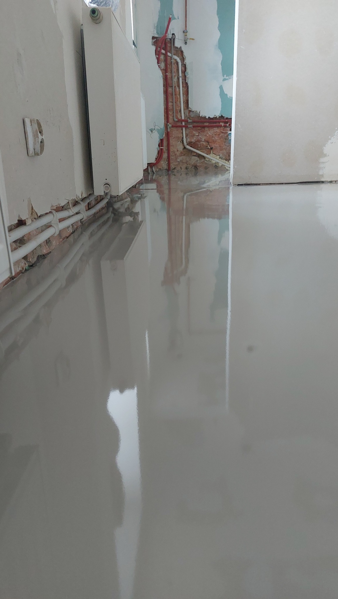 Świeżo wylana, gładka, szara posadzka żywiczna w pomieszczeniu w trakcie remontu. Widoczne instalacje grzewcze i fragmenty odsłoniętej cegły na ścianach.
