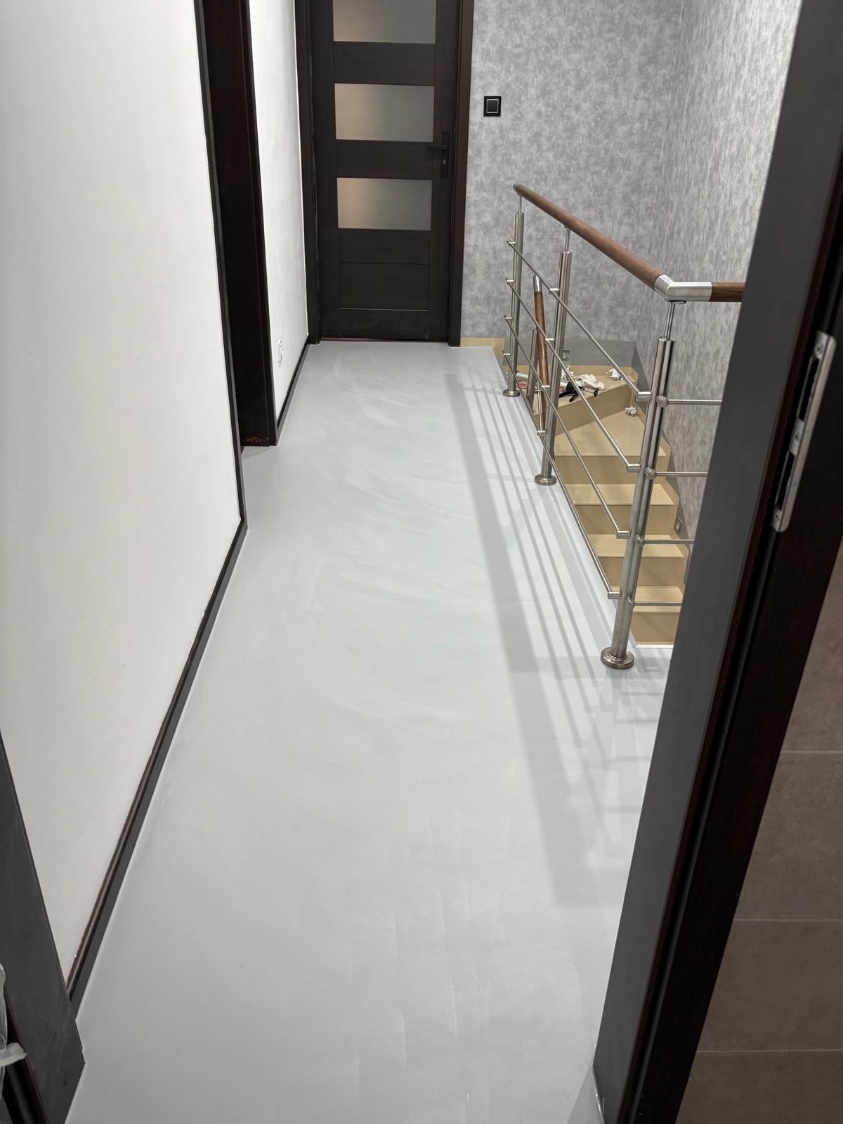 Jasnoszara, gładka posadzka w korytarzu z drewnianymi drzwiami i schodami z metalową balustradą. Ściany w odcieniach szarości. Widok z perspektywy wejścia.