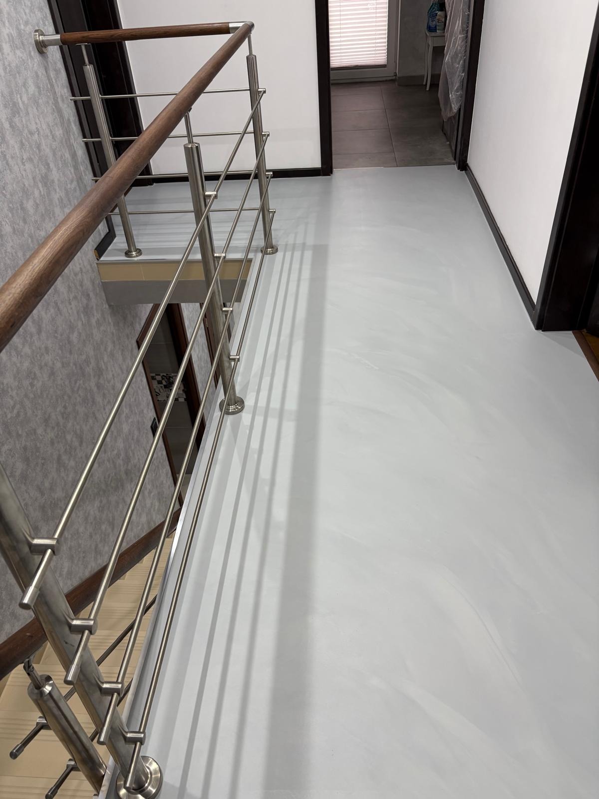 Jasnoszara, gładka posadzka żywiczna w korytarzu, widok z góry na schody z metalową balustradą i drewnianym poręczą. Minimalistyczne wnętrze z białymi ścianami.