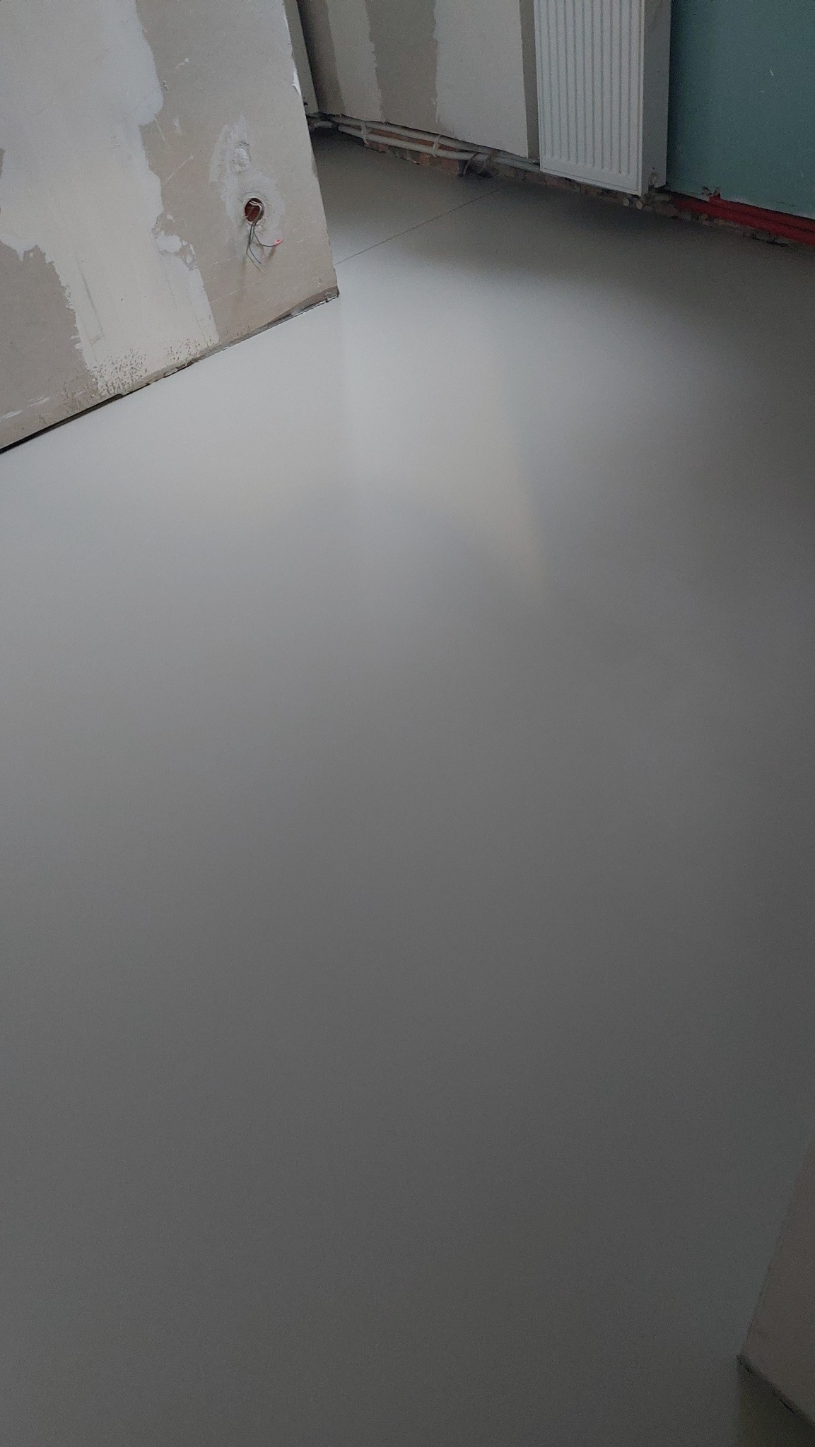 Gładka, szara posadzka w surowym wnętrzu z widocznymi przewodami elektrycznymi i grzejnikiem. Minimalistyczny design, świeżo wylana powierzchnia.