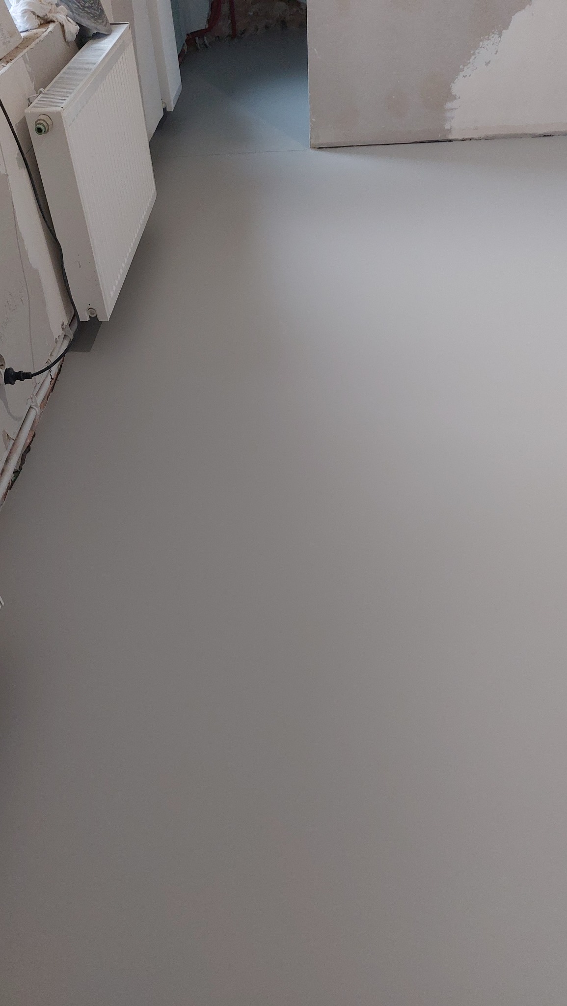 Gładka, szara posadzka w trakcie remontu, widoczny grzejnik i fragment ściany z niedokończonymi pracami tynkarskimi. Minimalistyczne ujęcie na podłogę.