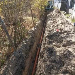 Wykop z ułożonym pomarańczowym rurociągiem, widoczna mini koparka na tle budynku i drzew. Świeżo usypana ziemia wzdłuż wykopu.