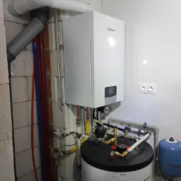 Nowoczesny, biały piec gazowy Vaillant z podłączonym zasobnikiem ciepłej wody i instalacją hydrauliczną w pomieszczeniu z białymi ścianami i widocznymi rurami wentylacyjnymi.