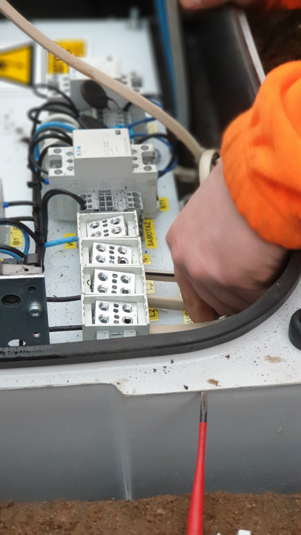 Ręka w pomarańczowej bluzie podłącza przewód do listwy zaciskowej w skrzynce elektrycznej; widoczny fragment czerwonego śrubokrętu.