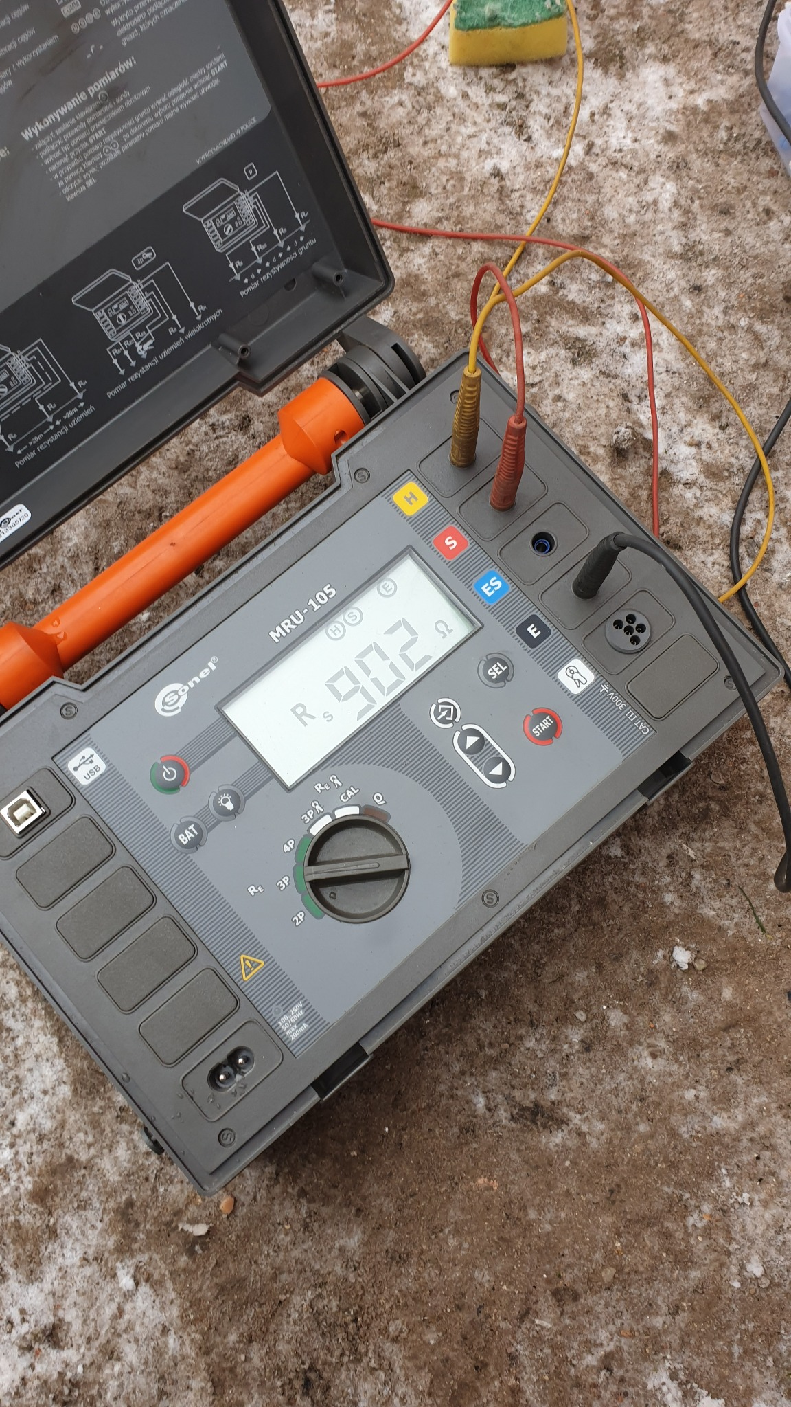 Miernik rezystancji uziemienia Sonel MRU-105 z podłączonymi przewodami pomiarowymi, wyświetlający wynik pomiaru na tle ziemi z resztkami śniegu.