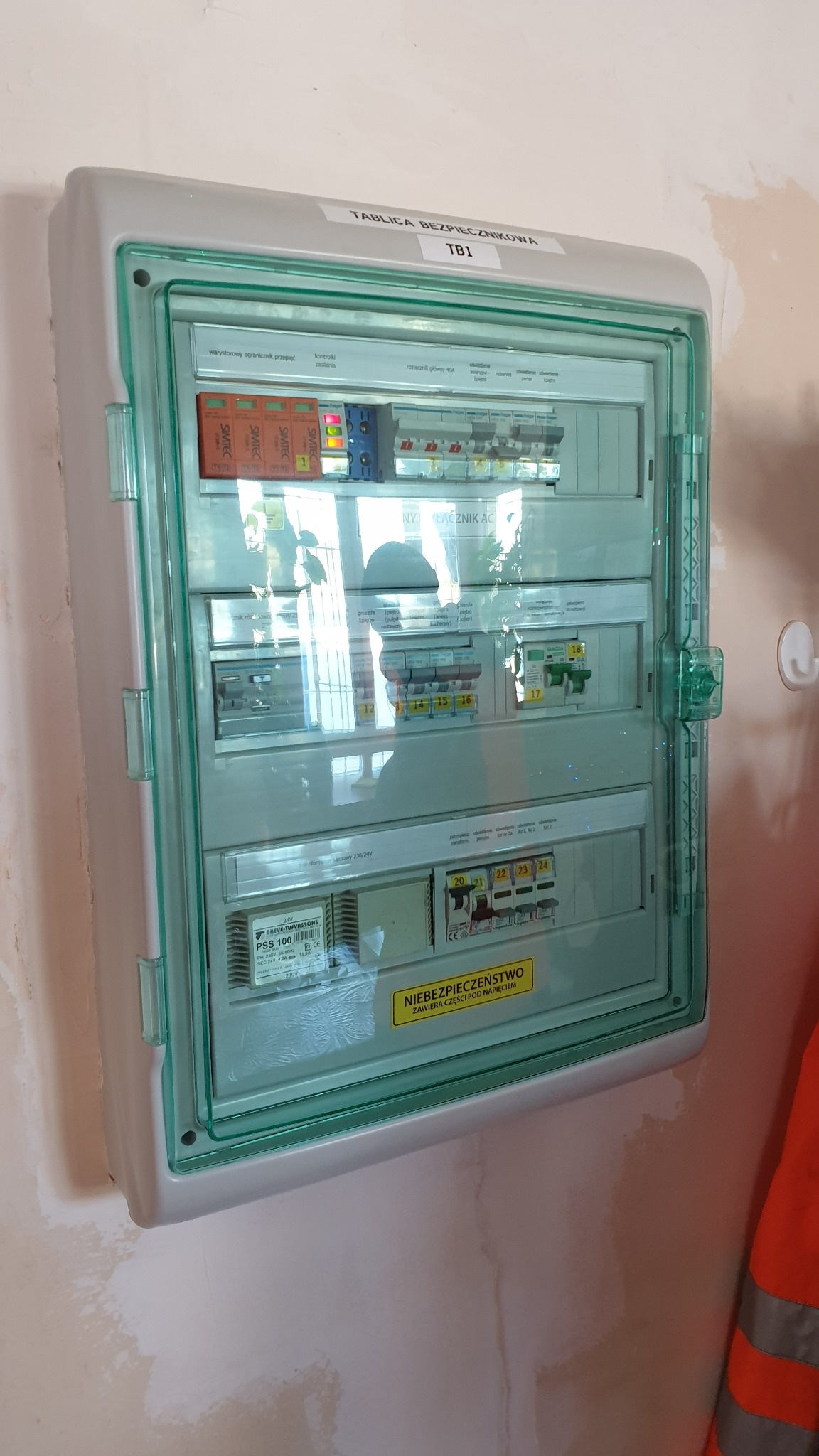Zamknięta tablica bezpiecznikowa TB1 z warystorami przepięć, kontrolkami zasilania i wyłącznikami, zamontowana na ścianie z widocznymi śladami po remoncie.