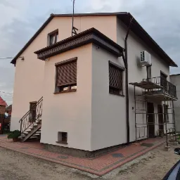 Dwupiętrowy dom z beżową elewacją, brązowymi okiennicami i widoczną jednostką zewnętrzną klimatyzacji na ścianie, z metalowym rusztowaniem opartym o balkon.