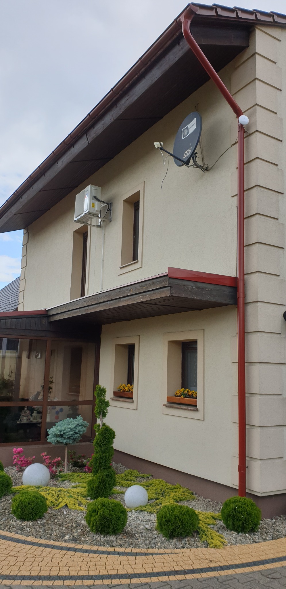 Zewnętrzna jednostka klimatyzacji zamontowana na ścianie domu, widoczne rurki odprowadzające skropliny, obok okna z brązową ramą, na elewacji beżowa farba.