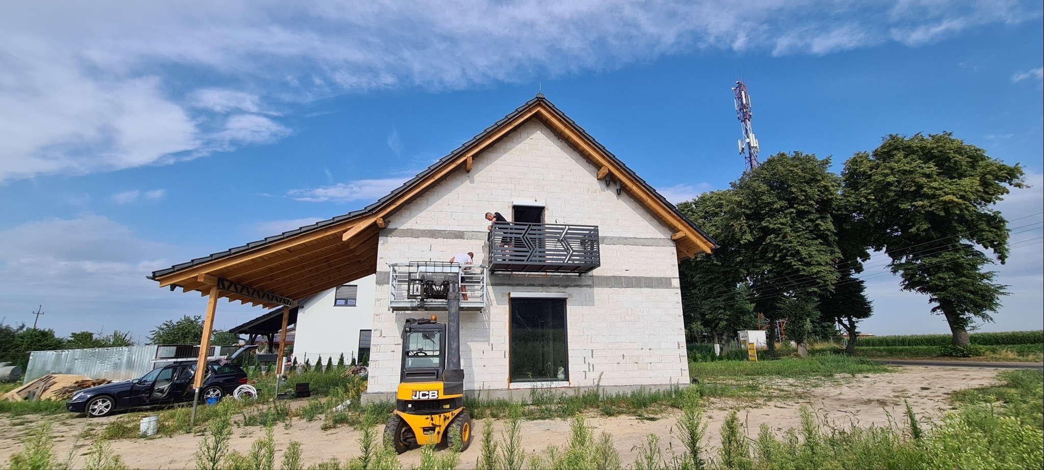Montaż balustrady na balkonie domu w trakcie budowy, z użyciem podnośnika koszowego JCB na tle błękitnego nieba