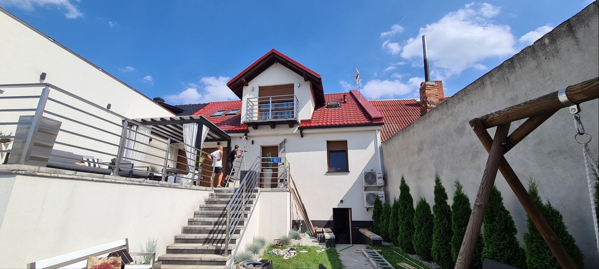 Montaż zewnętrznych jednostek klimatyzacji na elewacji domu z czerwonym dachem, widoczne dwie osoby na drabinie.