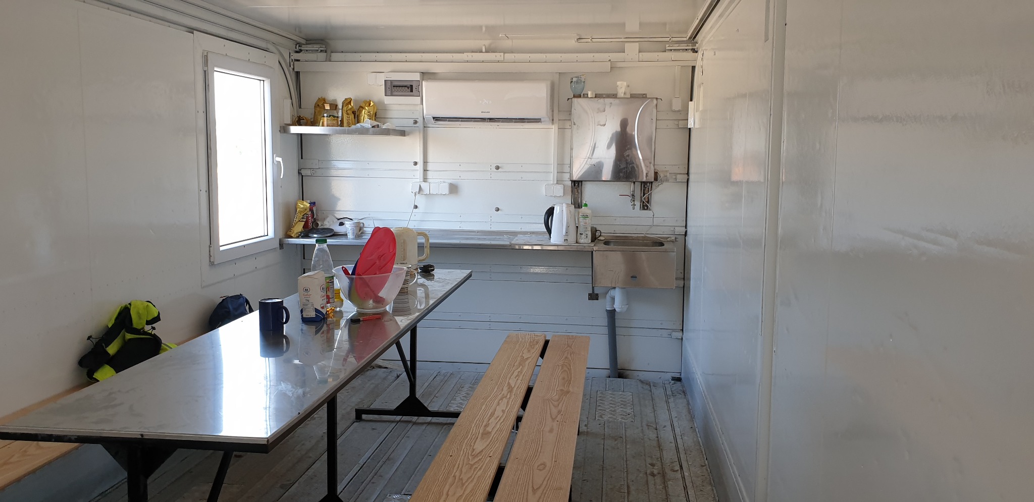 Wnętrze kontenera biurowego z zamontowaną klimatyzacją ścienną, wyposażonego w stół, ławki, zlew i półki z artykułami spożywczymi.