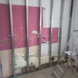 Metalowy stelaż pod płytę gipsową, częściowo wypełniony pianką montażową, widoczna instalacja hydrauliczna, fragment różowej ściany i drabina malarska.