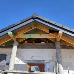 Wznoszenie dachu domu jednorodzinnego z szarą dachówką ceramiczną i drewnianą konstrukcją więźby, widoczne słupy wsporcze i ściana z bloczków betonu komórkowego na tle błękitnego nieba.