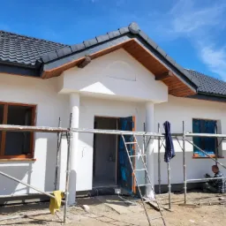 Pracownik siedzi na ziemi i szpachluje podmurówkę domu z białą elewacją na tle błękitnego nieba, widoczne rusztowania i zabezpieczone okna folią.