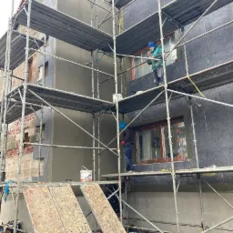 Budynek w trakcie ocieplania: fragment elewacji z szarym styropianem, rusztowanie z pracownikami, widoczna część budynku przed ociepleniem z surową cegłą i tynkiem.