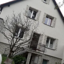 Dwupiętrowy dom z jasną elewacją, ozdobiony kamiennymi panelami wokół okien i na cokole, z balkonem i drzewem bez liści na pierwszym planie.