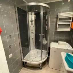 Narożna kabina prysznicowa z hydromasażem, umieszczona w łazience z szarymi płytkami, obok pralki i umywalki z niebieską miską.