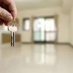 Klucz do mieszkania trzymany w dłoni na tle jasnego, pustego pokoju z błyszczącą podłogą i oknem balkonowym.