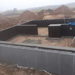 Fundamenty domu w trakcie budowy, z izolacją przeciwwilgociową, widoczne wyjścia na instalacje, w tle hałdy ziemi i zalesiony teren w porannej mgle.