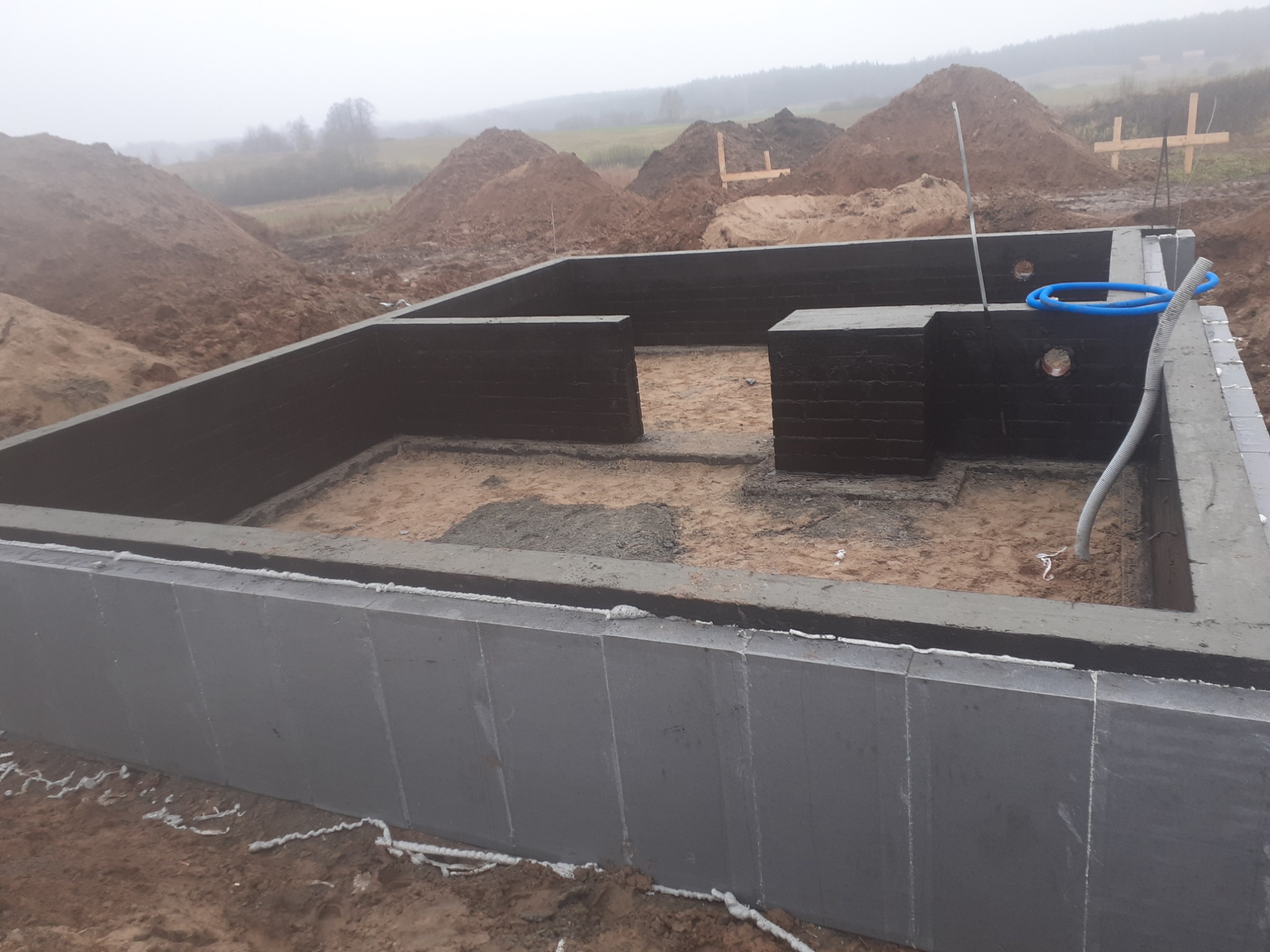 Fundamenty domu w trakcie budowy, z izolacją przeciwwilgociową, widoczne wyjścia na instalacje, w tle hałdy ziemi i zalesiony teren w porannej mgle.
