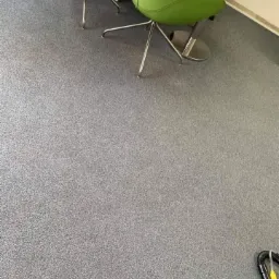 Szary dywan w biurze po czyszczeniu, widoczne fragmenty zielonych krzeseł i okrągłego stolika, w rogu fragment odkurzacza piorącego.
