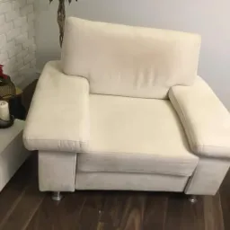 Jasny, beżowy fotel tapicerowany z podłokietnikami, ustawiony na ciemnej drewnianej podłodze, z widocznymi metalowymi nóżkami i rośliną poinsecji w tle.