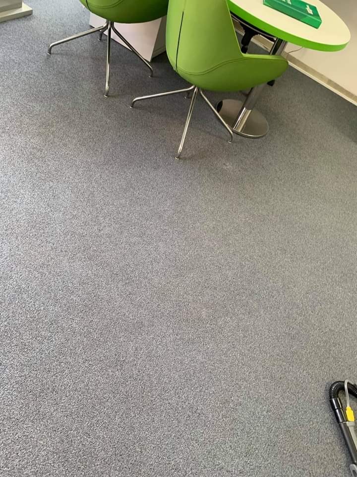 Szary dywan w biurze po czyszczeniu, widoczne fragmenty zielonych krzeseł i okrągłego stolika, w rogu fragment odkurzacza piorącego.